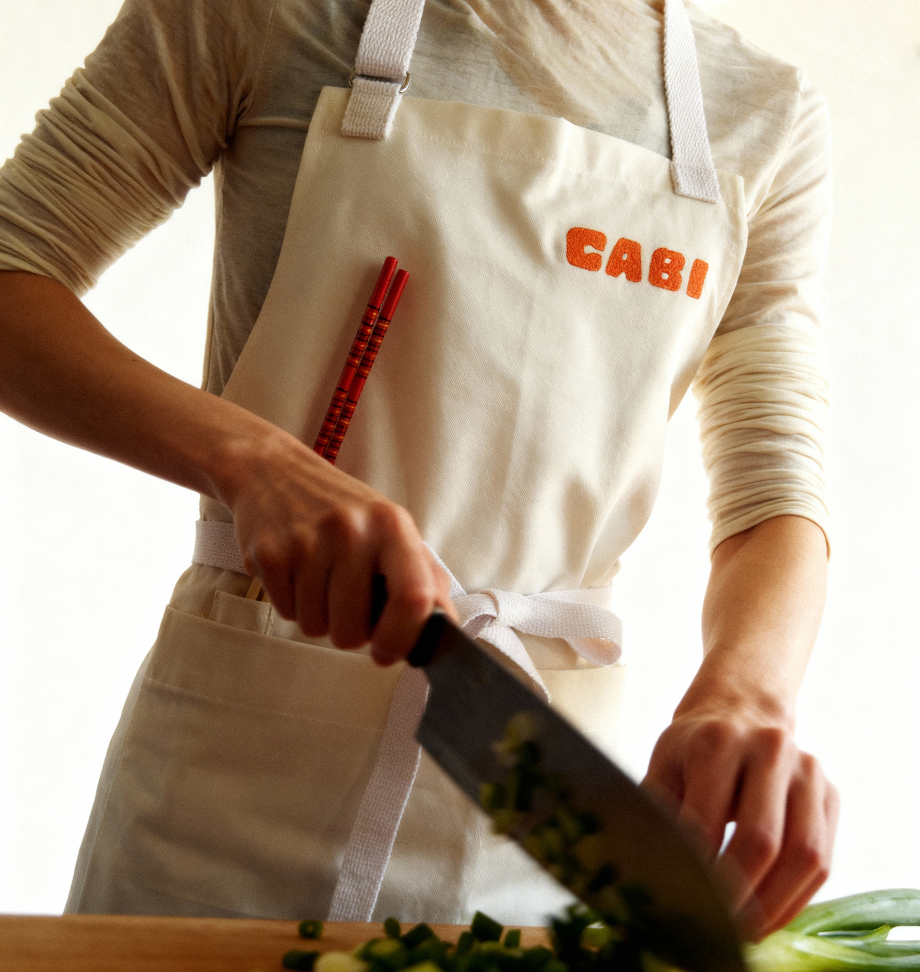 Person wearing a 'Cabi' apron, holding a knife and cutting vegetables on a wooden board.