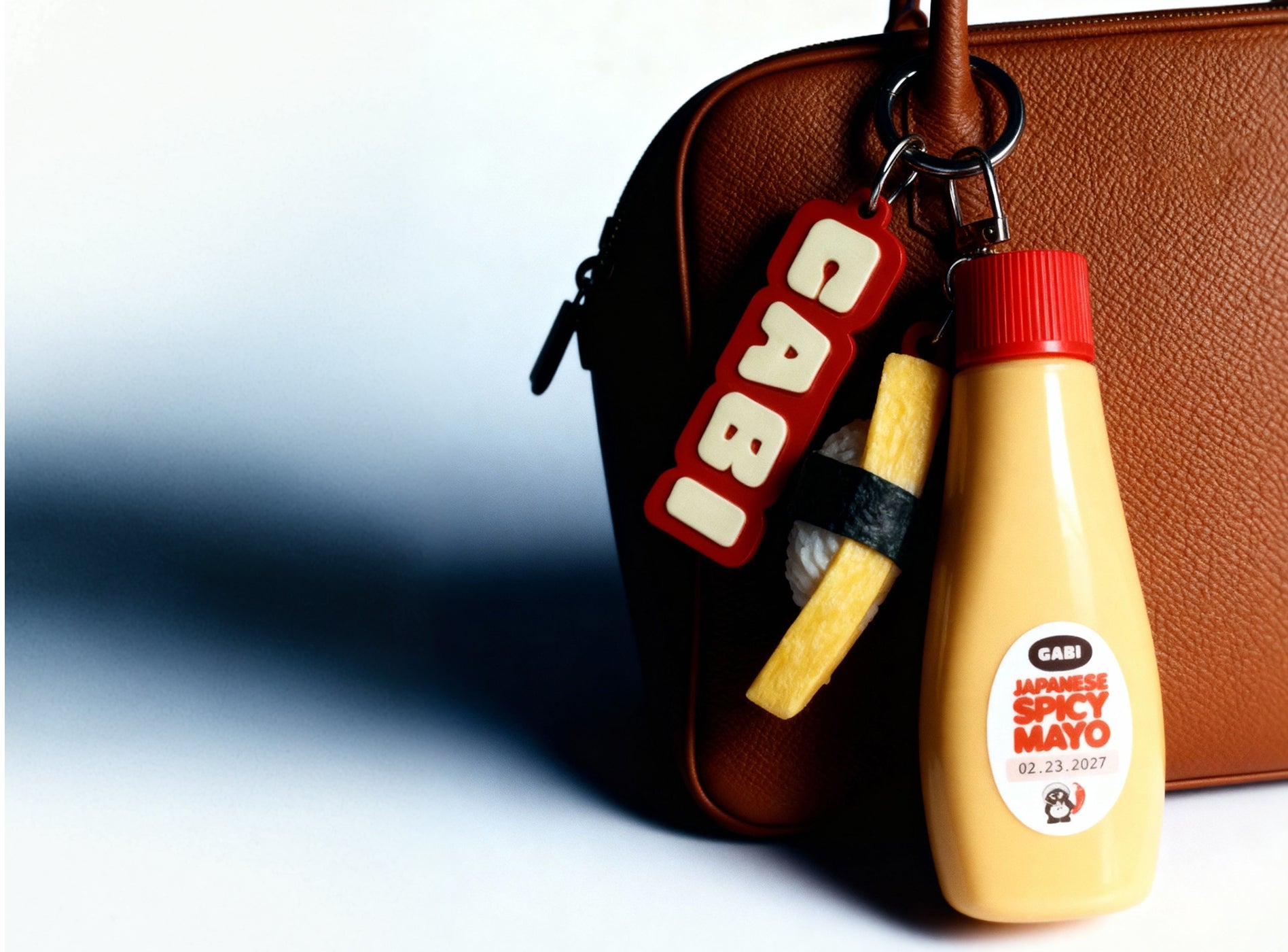 Keychain shaped like a Cabi mayo bottle with a red cap on a brown leather bag.