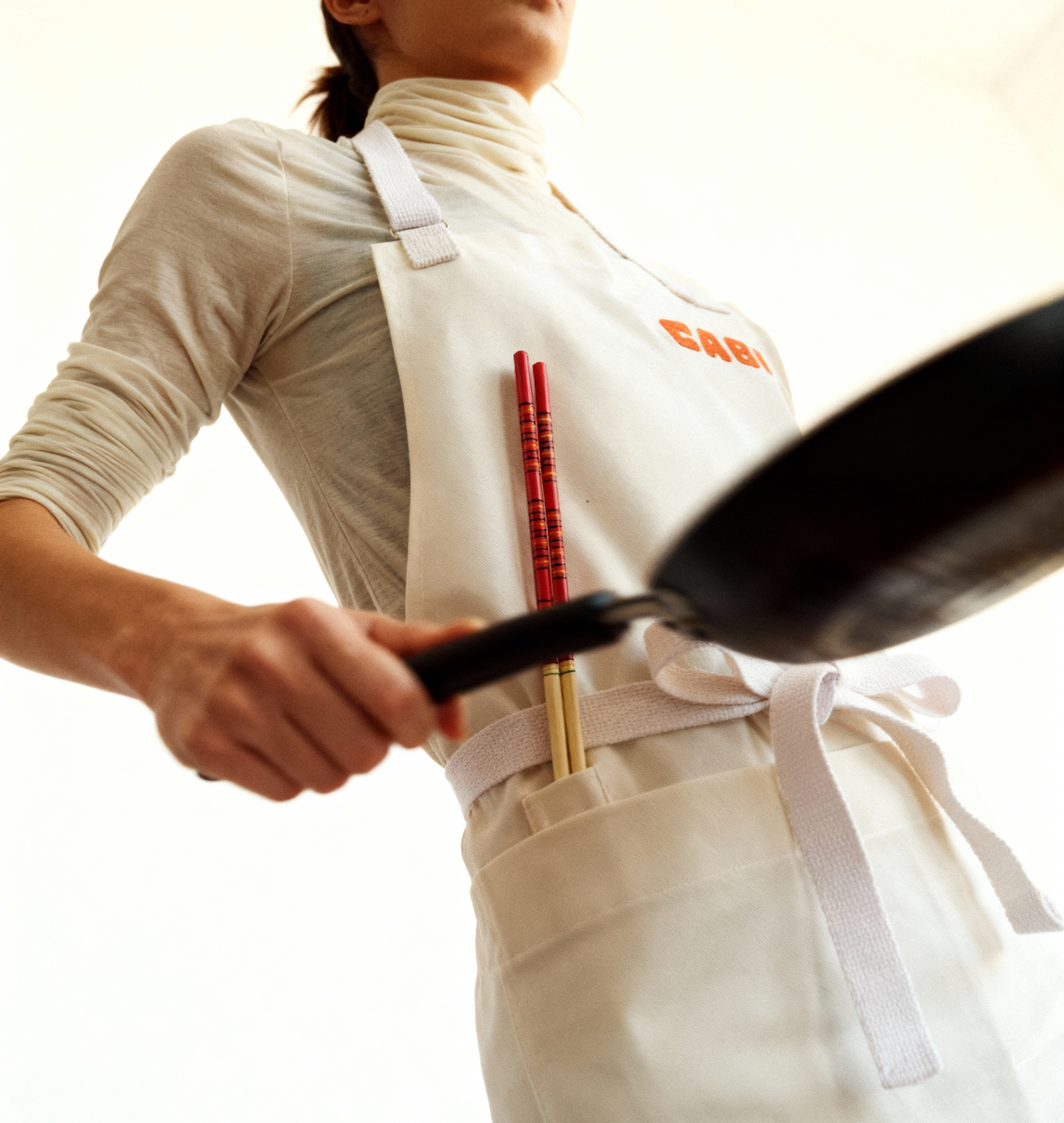 Person wearing a white apron with 'Cabi' text, holding a frying pan.
