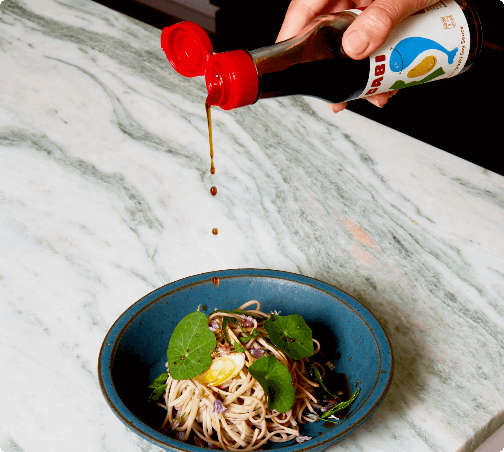 Cabi Umami Dashi Soy Sauce being drizzled over soba noodles with soft-boiled egg and fresh greens in a blue bowl.