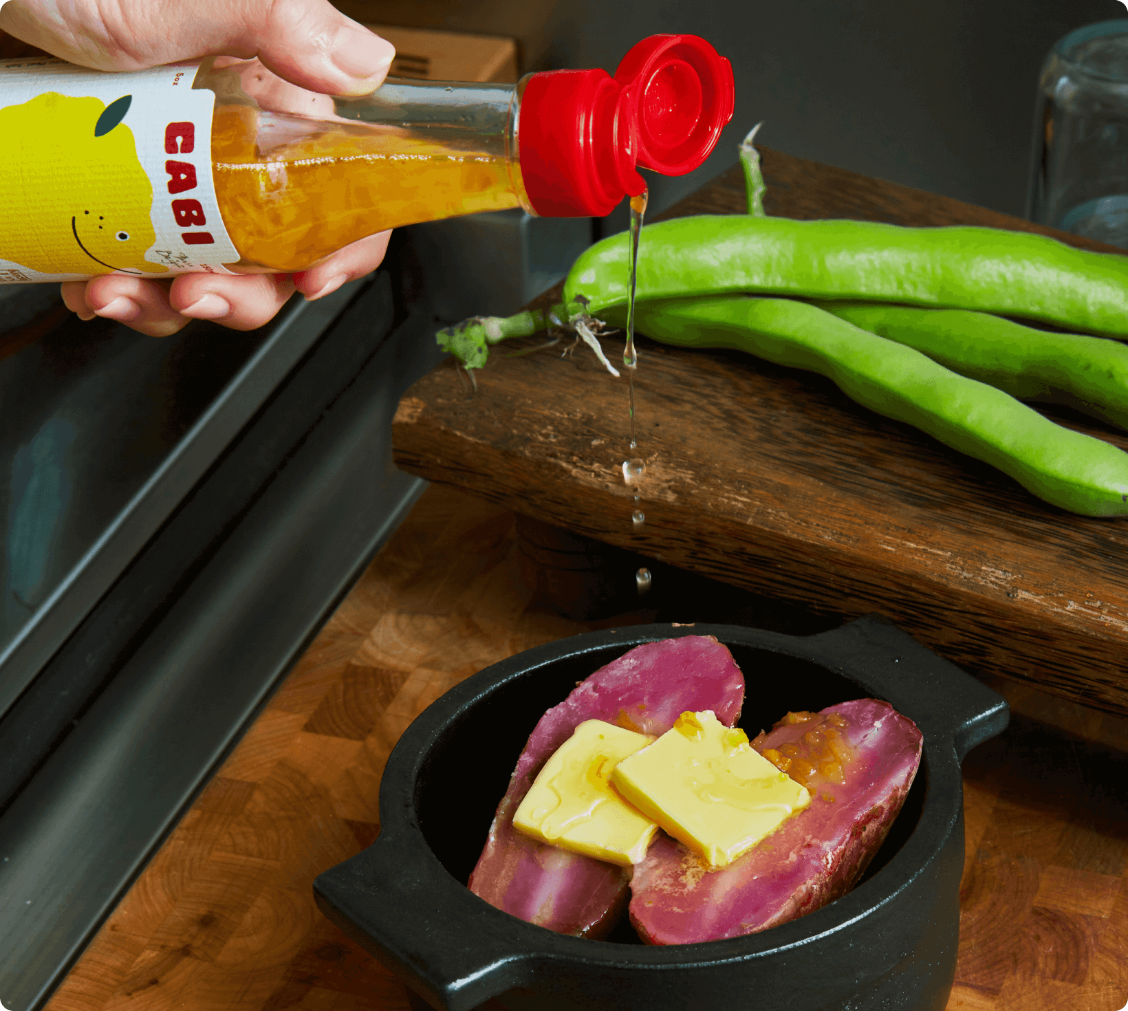 Cabi Sweet Yuzu Vinaigrette drizzled over Sweet Potatoe with butter, with fresh green peppers on a wooden cutting board.
